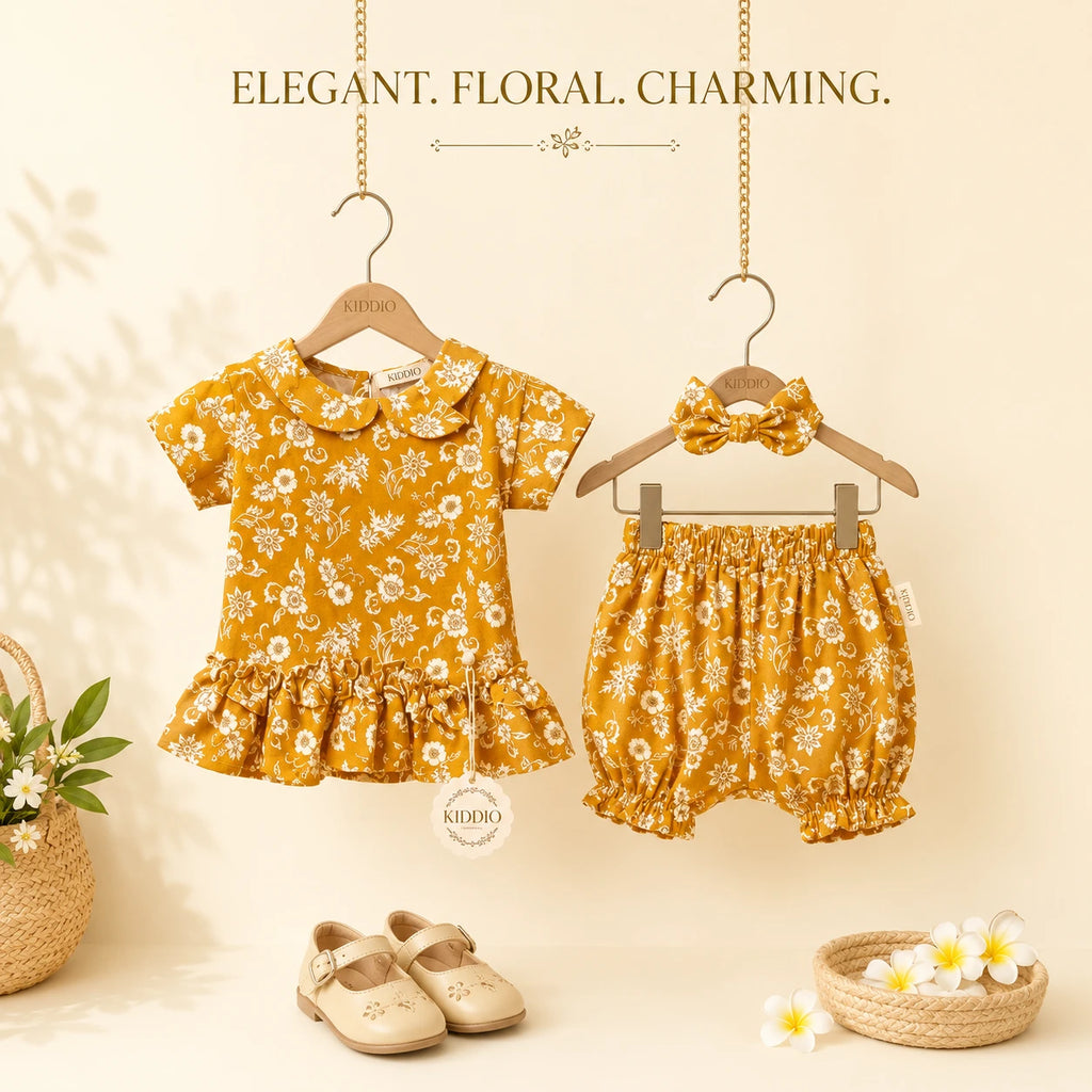 A baby girl's two-piece outfit set featuring a short-sleeve top and matching bloomers, hanging on wooden hangers. The outfit is a mustard yellow color with a white floral print.