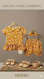 A coordinated baby outfit set displayed on wooden hangers against a warm taupe background. The set includes a mustard yellow floral short-sleeve top with a ruffled hem and matching floral bloomers, plus a matching bow headband.