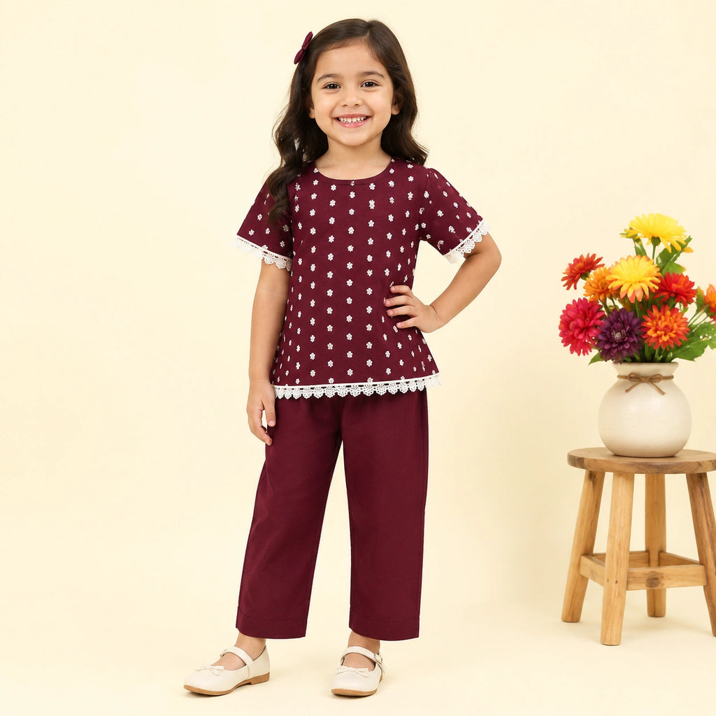 A young girl modeling a two-piece outfit consisting of a maroon short-sleeve top with white polka dots and white lace trim, paired with matching maroon cropped pants. She is standing next to a small wooden stool holding a vase of colorful flowers against a solid light beige background.