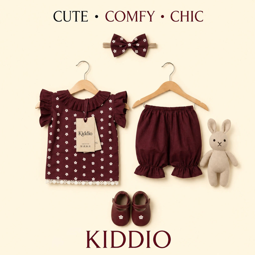 A baby girl's outfit set on a cream background, featuring a dark burgundy short-sleeve top with white daisy print and ruffles, matching bloomers, a bow headband, a plush bunny toy, and baby shoes. The brand name 'KIDDIO' is printed at both the top and bottom.