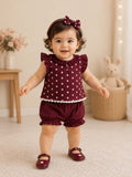 A baby girl wearing a burgundy two-piece outfit with white floral polka dots, ruffled sleeves, and matching ruffled shorts, along with a matching bow headband and burgundy shoes, standing in a cozy room.