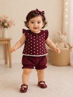 A baby girl wearing a burgundy two-piece outfit with white floral polka dots, ruffled sleeves, and matching ruffled shorts, along with a matching bow headband and burgundy shoes, standing in a cozy room.