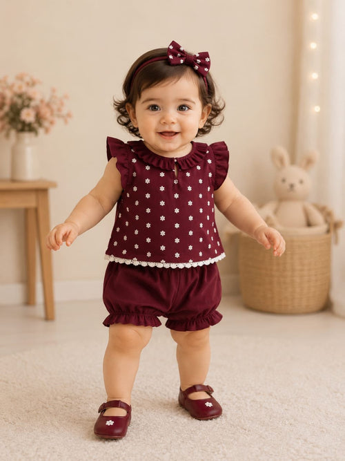 A baby girl wearing a burgundy two-piece outfit with white floral polka dots, ruffled sleeves, and matching ruffled shorts, along with a matching bow headband and burgundy shoes, standing in a cozy room.