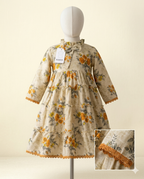 A cream-colored toddler dress with a yellow and green floral print, displayed on a mannequin. The dress features a tiered skirt, three-quarter sleeves, and a ruffled collar with a bow.