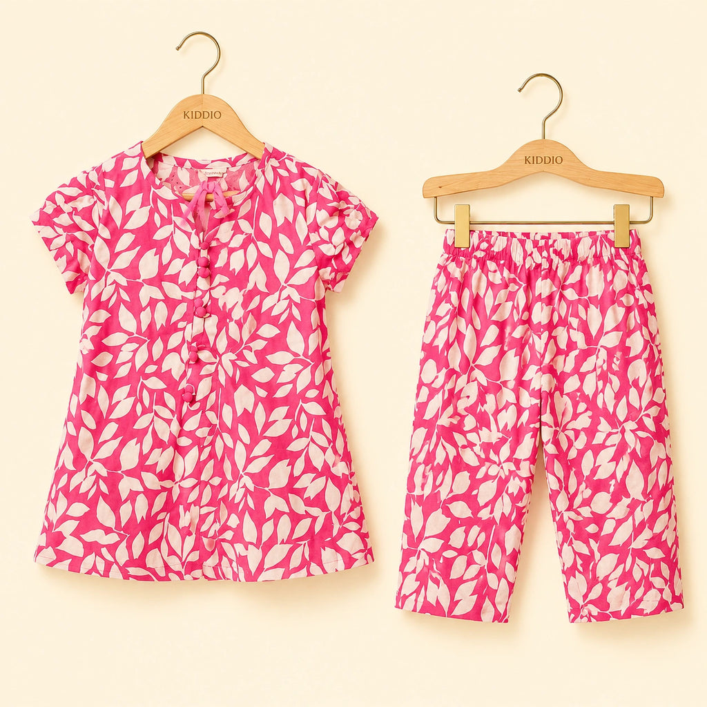 A two-piece children's outfit set consisting of a short-sleeve button-front top and matching cropped pants, both in a bright pink and white leaf pattern, hanging on wooden KIDDIO hangers against a light cream background.