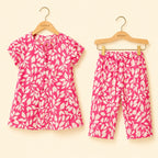 A two-piece children's outfit set consisting of a short-sleeve button-front top and matching cropped pants, both in a bright pink and white leaf pattern, hanging on wooden KIDDIO hangers against a light cream background.