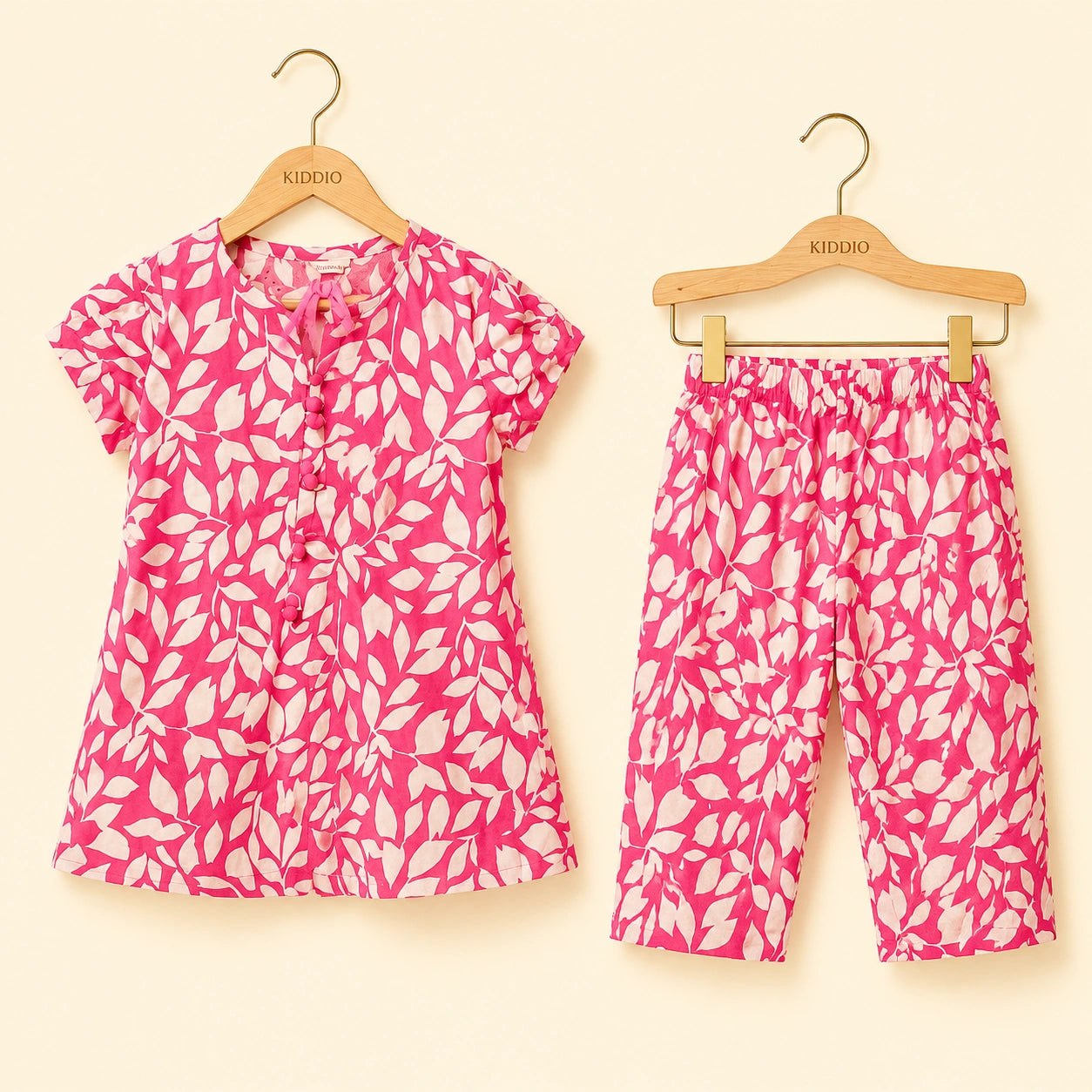 A two-piece children's outfit set consisting of a short-sleeve button-front top and matching cropped pants, both in a bright pink and white leaf pattern, hanging on wooden KIDDIO hangers against a light cream background.