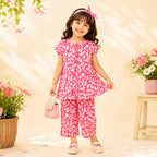 A young girl stands smiling in a pink and white patterned two-piece outfit with a matching headband, holding a small pink handbag. The scene is styled with flowers, a basket, and a small table against a warm cream backdrop.
