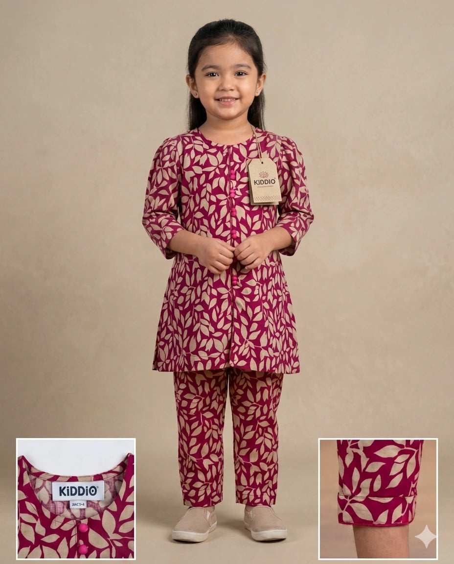 A young girl stands against a plain beige studio background wearing a matching two-piece outfit with a magenta and cream leaf pattern. The set includes a long-sleeve tunic and pants.
