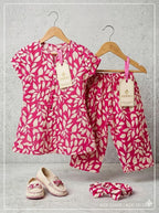 A two-piece children's outfit set on wooden hangers, featuring a short-sleeve top and matching pants in a bright pink and cream leaf pattern. A matching fabric headband and a pair of cream slip-on shoes with pink floral details are displayed below on a textured gray background.