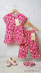 A children's two-piece outfit set hanging on wooden hangers against a light gray wall, featuring a short-sleeve top and matching pants in bright pink and cream with a leaf print. A pair of cream slip-on shoes and a matching cream headband are placed below.