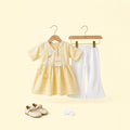 A baby girl's two-piece outfit set on wooden hangers, featuring a pale yellow short-sleeve dress with white lace trim and white bell-bottom pants. A pair of cream-colored baby shoes and a matching white bow are placed below the outfit against a solid pale yellow background.