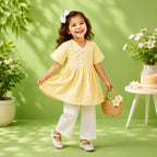 A young girl smiling and twirling in a pale yellow embroidered dress with white pants, holding a small woven basket with daisy flowers, posed in a bright green studio setting with potted flowers.