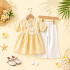 A baby girl's two-piece outfit set featuring a pale yellow short-sleeve dress with lace trim and a white pair of wide-leg pants, displayed on wooden hangers with decorative props like a straw hat, flowers, and a palm leaf.