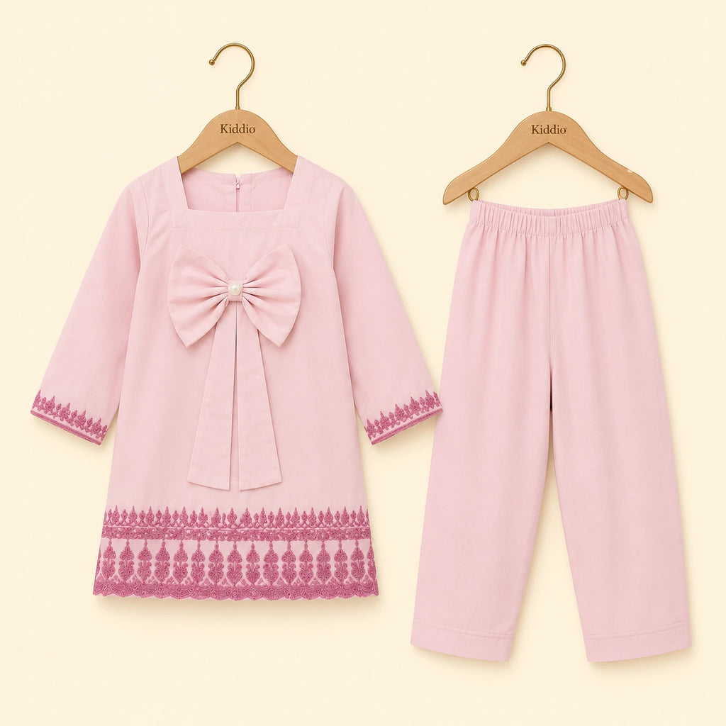 A two-piece baby girl's outfit set consisting of a light pink long-sleeve dress with a large bow on the chest and a pink lace hem, paired with matching light pink pants. Both pieces are displayed on wooden hangers with the brand name 'Kiddo' printed on them, set against a solid pale cream background.
