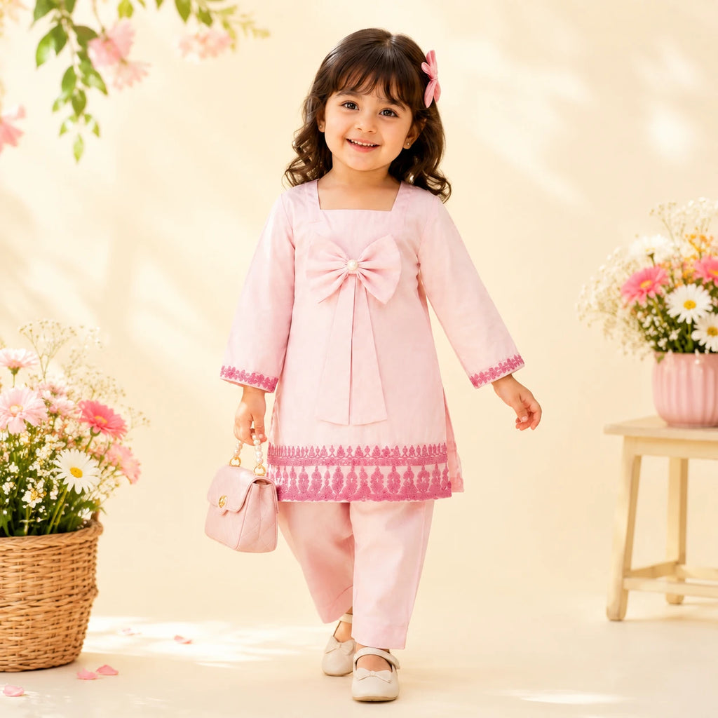 A young girl wearing a light pink traditional outfit with a large bow at the chest, matching pink pants, and a small pink handbag, standing in a warm, sunlit studio setting with flowers and soft shadows.