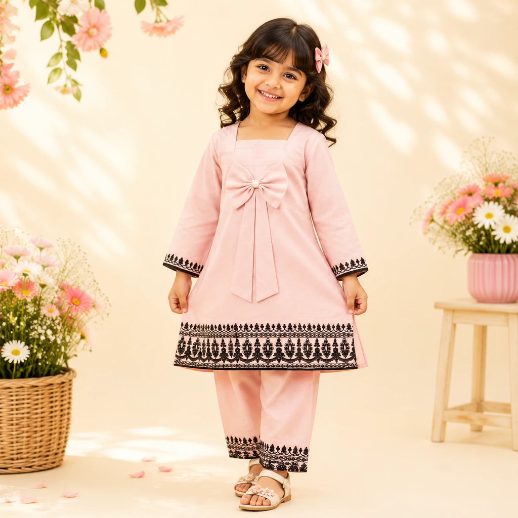 A young girl modeling a light pink traditional outfit with black embroidered borders, standing in a warm, decorated studio setting with flowers.