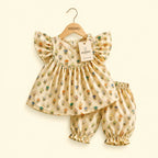 A baby girl's two-piece outfit set consisting of a flutter-sleeve dress and matching bloomers, hanging on a wooden KIDDIO hanger against a warm cream background. The outfit is cream-colored with a colorful pattern of cartoon characters, and features a KIDDIO brand tag.