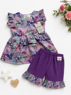 A toddler girl's two-piece outfit set from Koodio, featuring a purple and teal floral print ruffle dress and solid purple ruffle-trimmed shorts, styled on a white background with decorative flowers and star props.