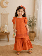 A young girl stands in a styled indoor room wearing a bright orange two-piece outfit with puff sleeves, a scalloped lace hem, and wide flared pants. She also wears a matching orange bow headband, brown sandals, and carries a small woven crossbody bag.