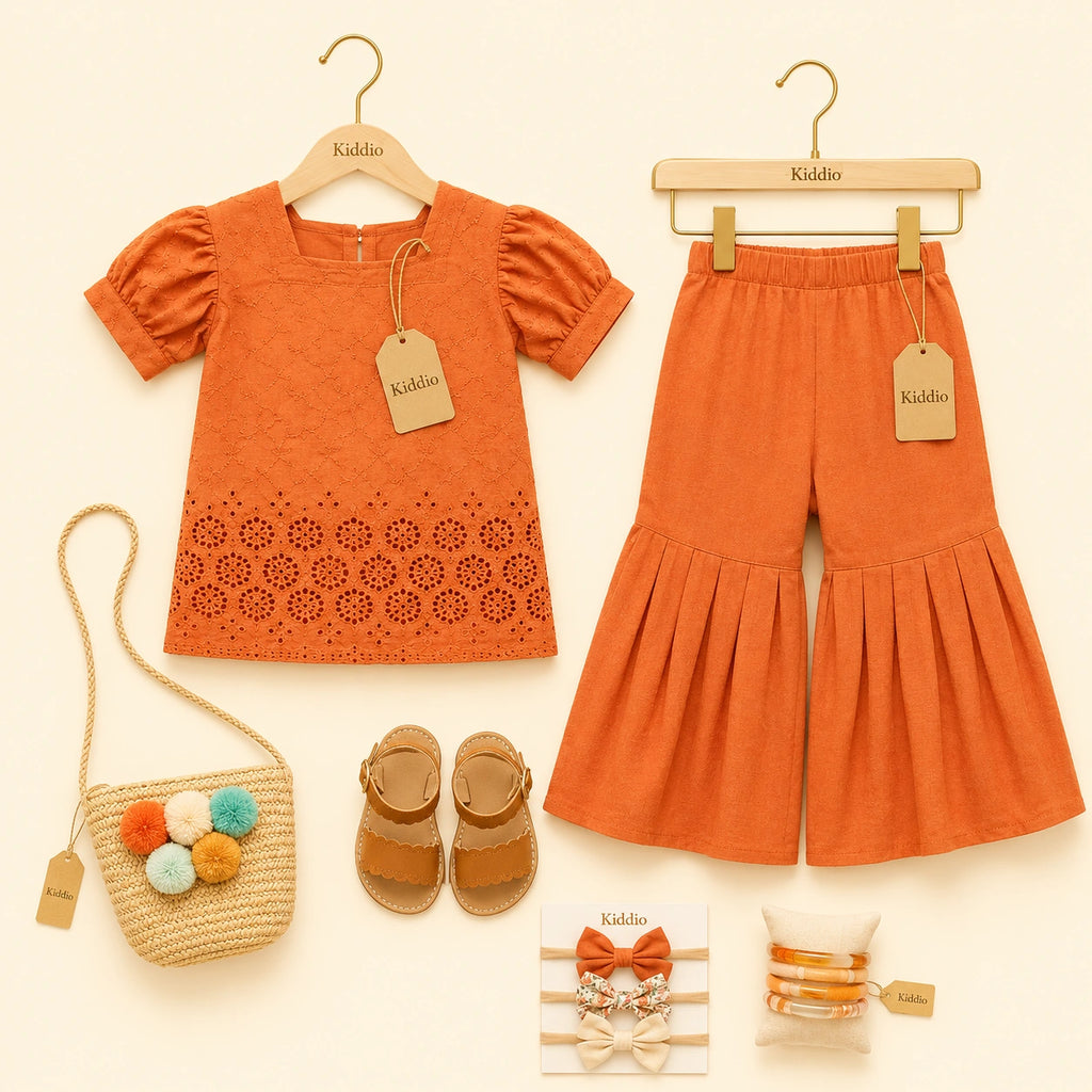 A children's outfit set displayed on a cream background, including an orange short-sleeve top and matching orange flared pants on wooden hangers, a woven straw bag with pom-poms, tan sandals, and accessories with Kiddio branding.