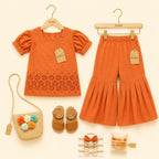 A children's outfit set displayed on a cream background, including an orange short-sleeve top and matching orange flared pants on wooden hangers, a woven straw bag with pom-poms, tan sandals, and accessories with Kiddio branding.