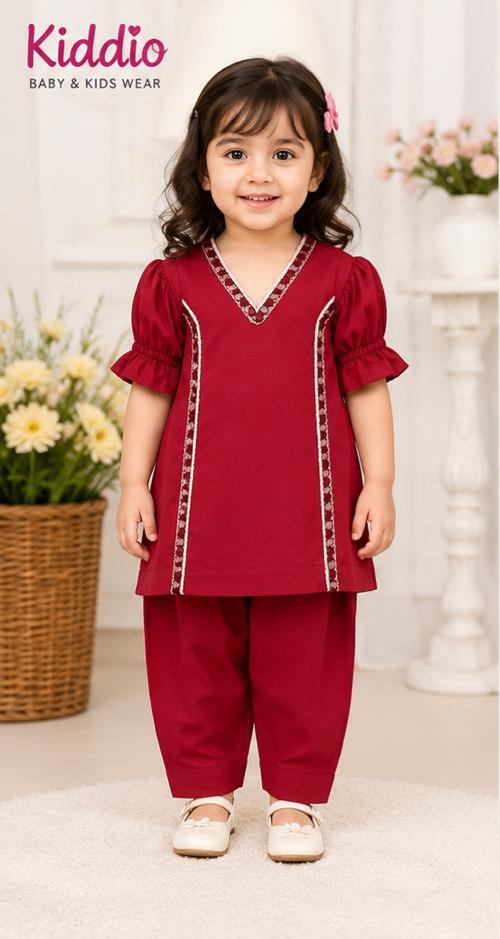 A young girl modeling a deep red two-piece traditional outfit with white and gold embroidered details, standing in a bright indoor setting with floral decorations. The brand logo 'Kiddio Baby & Kids Wear' is visible in the top left corner.