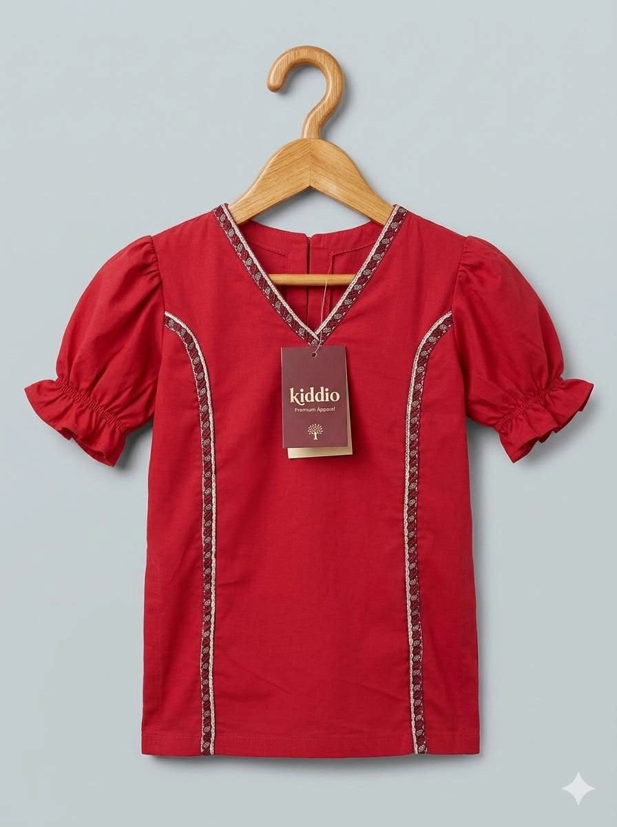 A red short-sleeve baby top with puff sleeves and decorative embroidered trim, hanging on a wooden hanger against a light gray background. A brand tag reading 'kiddio' is attached to the neckline.