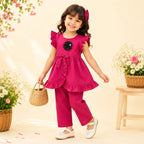 A young girl smiling and posing in a bright magenta pink two-piece outfit consisting of a ruffled peplum top with a black flower applique and matching wide-leg pants. She is holding a small woven basket bag and wearing white Mary Jane shoes, set against a warm cream background with decorative flowers.