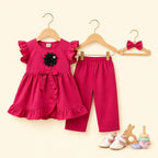 A baby girl's two-piece outfit set consisting of a bright pink ruffle-sleeve top with a black flower applique and matching pink pants, displayed on wooden hangers alongside a matching pink bow headband. The set is photographed against a solid pale cream background with small toy props at the bottom right.