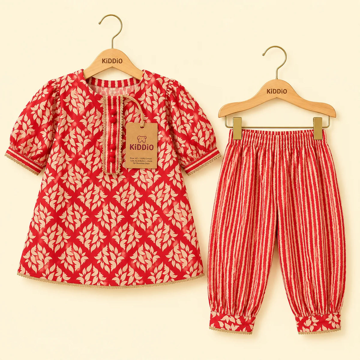 A two-piece children's outfit set by Kiddio, featuring a red and beige leaf-patterned short-sleeve top on a wooden hanger and matching red and beige vertically striped pants on a second hanger, displayed against a light cream background.