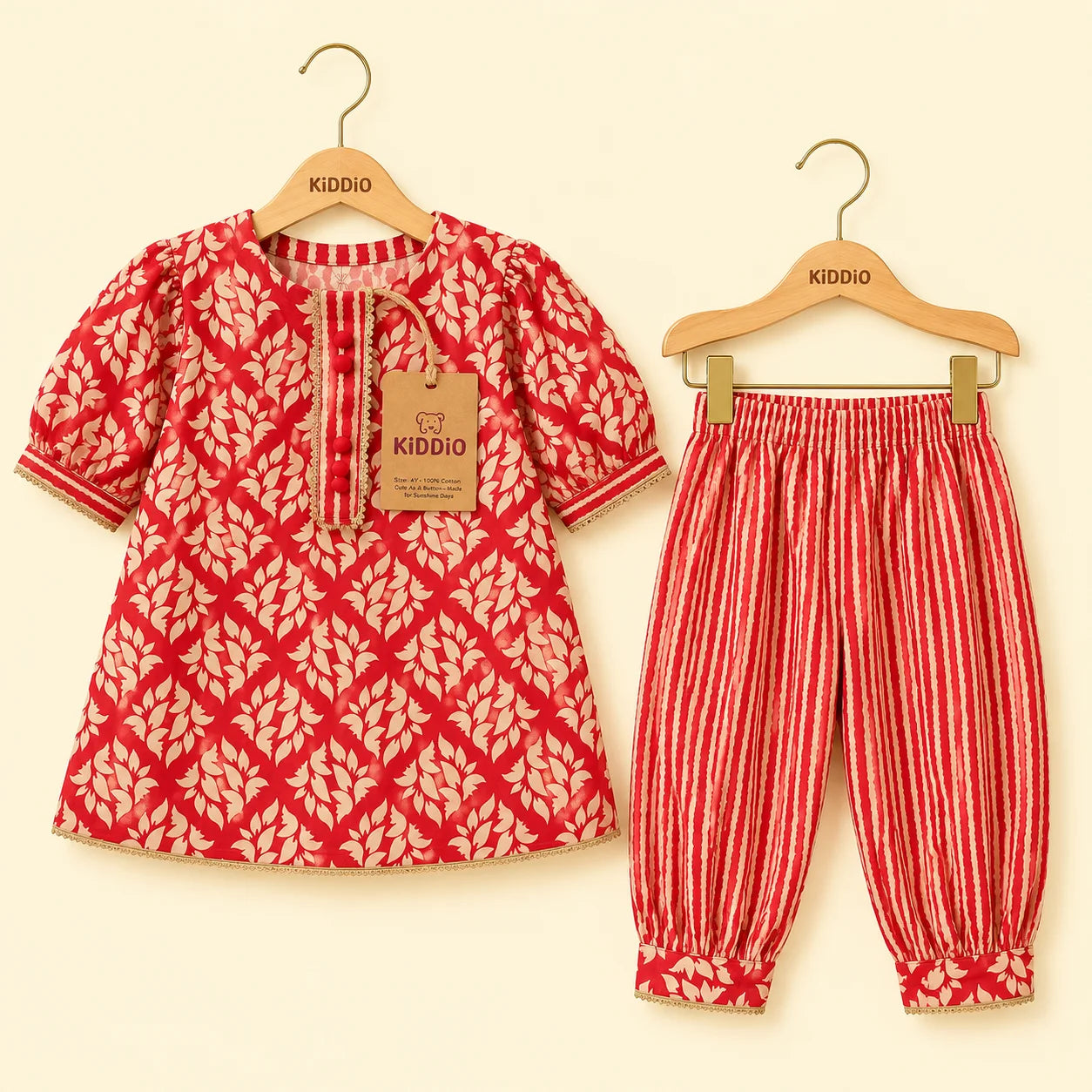 A two-piece children's outfit set by Kiddio, featuring a red and beige leaf-patterned short-sleeve top on a wooden hanger and matching red and beige vertically striped pants on a second hanger, displayed against a light cream background.