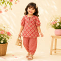 A young girl smiling and wearing a red and beige patterned kurta with matching red-and-beige striped pants, holding a small woven bag in a styled studio setting with flowers and a stool.