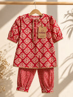 A children's two-piece outfit hanging on a wooden hanger against a warm beige wall with soft leaf shadows. The set includes a short-sleeve tunic top with a red and cream geometric leaf pattern and matching striped trim, plus striped pants with the same leaf motif.