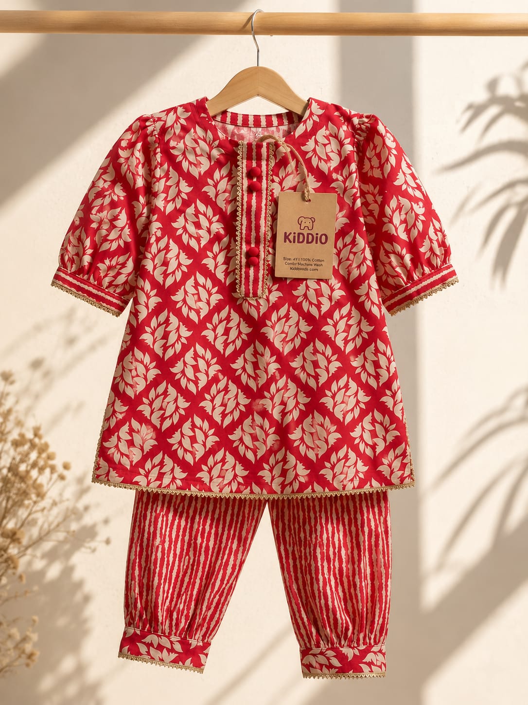 A children's two-piece outfit hanging on a wooden hanger against a warm beige wall with soft leaf shadows. The set includes a short-sleeve tunic top with a red and cream geometric leaf pattern and matching striped trim, plus striped pants with the same leaf motif.