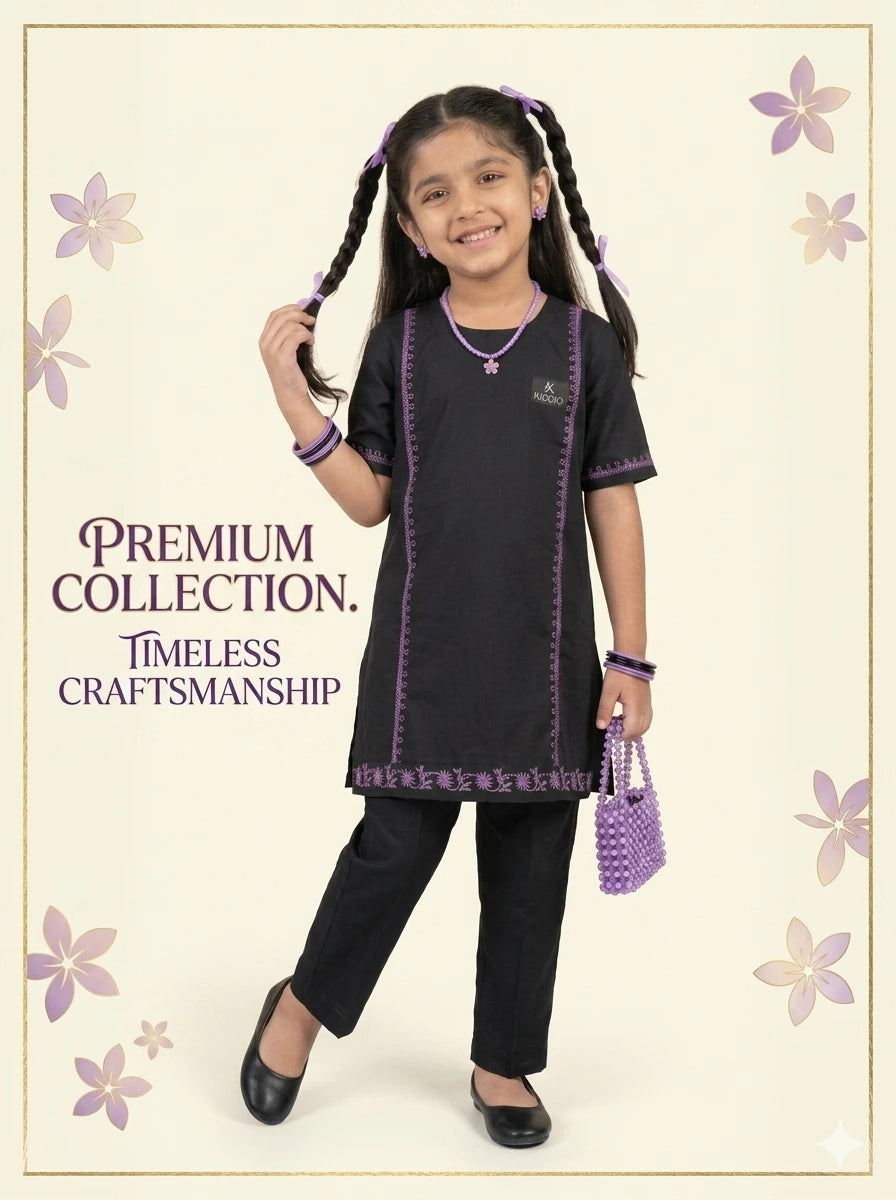 A young girl with braided pigtails models a black kurta and trouser set with purple embroidered borders. She is holding a small purple beaded handbag against a cream-colored background decorated with purple floral motifs and the text 'PREMIUM COLLECTION.