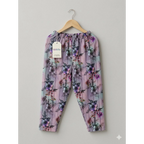 A pair of toddler girl's floral print pants in lavender and pink, hanging on a wooden hanger against a neutral gray background. The pants feature a purple and blue floral pattern over a light pink base, with an elastic waistband and a visible 'kiddio' brand tag.