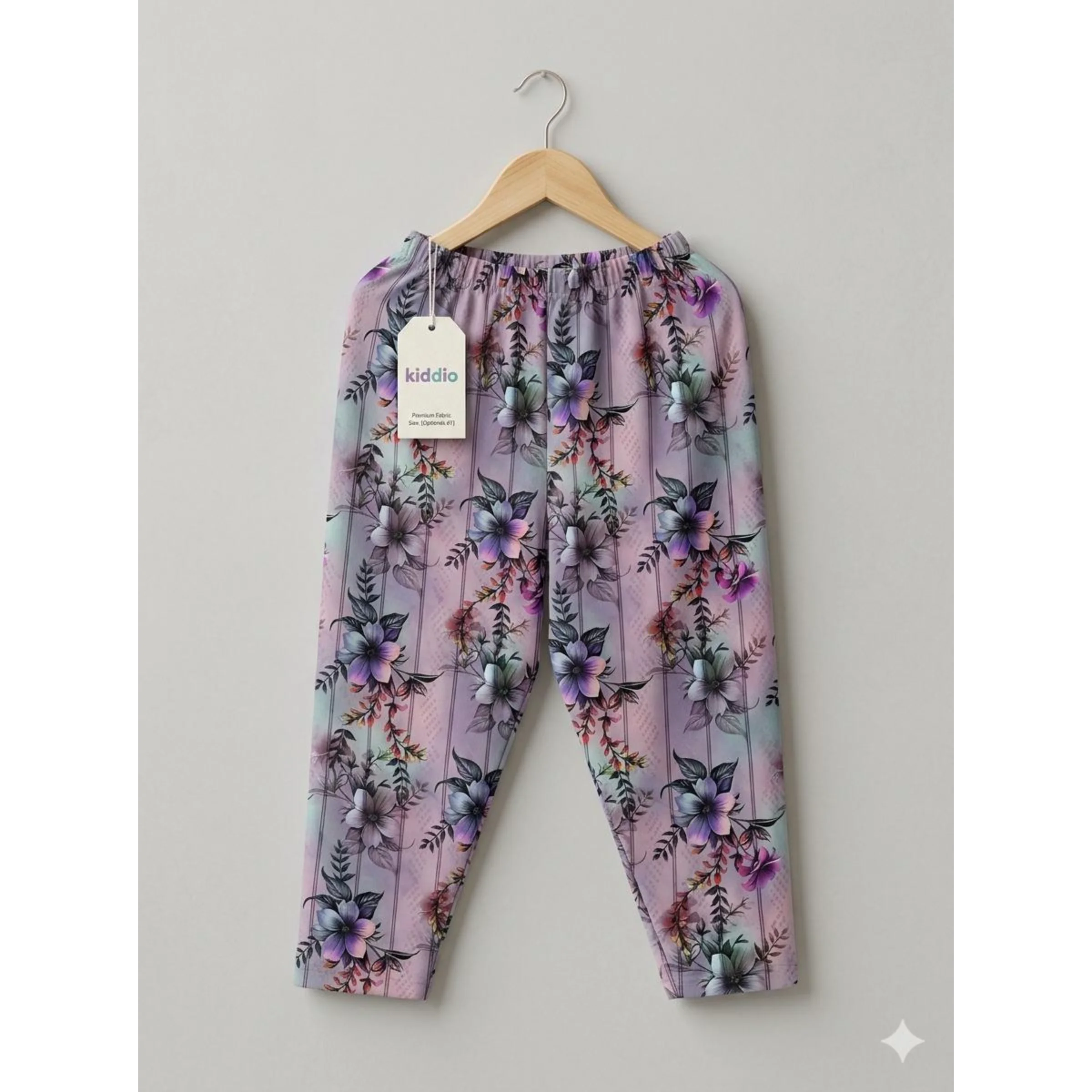 A pair of toddler girl's floral print pants in lavender and pink, hanging on a wooden hanger against a neutral gray background. The pants feature a purple and blue floral pattern over a light pink base, with an elastic waistband and a visible 'kiddio' brand tag.