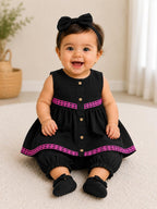 A baby girl sitting on a cream carpet wearing a black sleeveless romper with pink patterned trim, matching black pants, black socks, and a black bow headband.