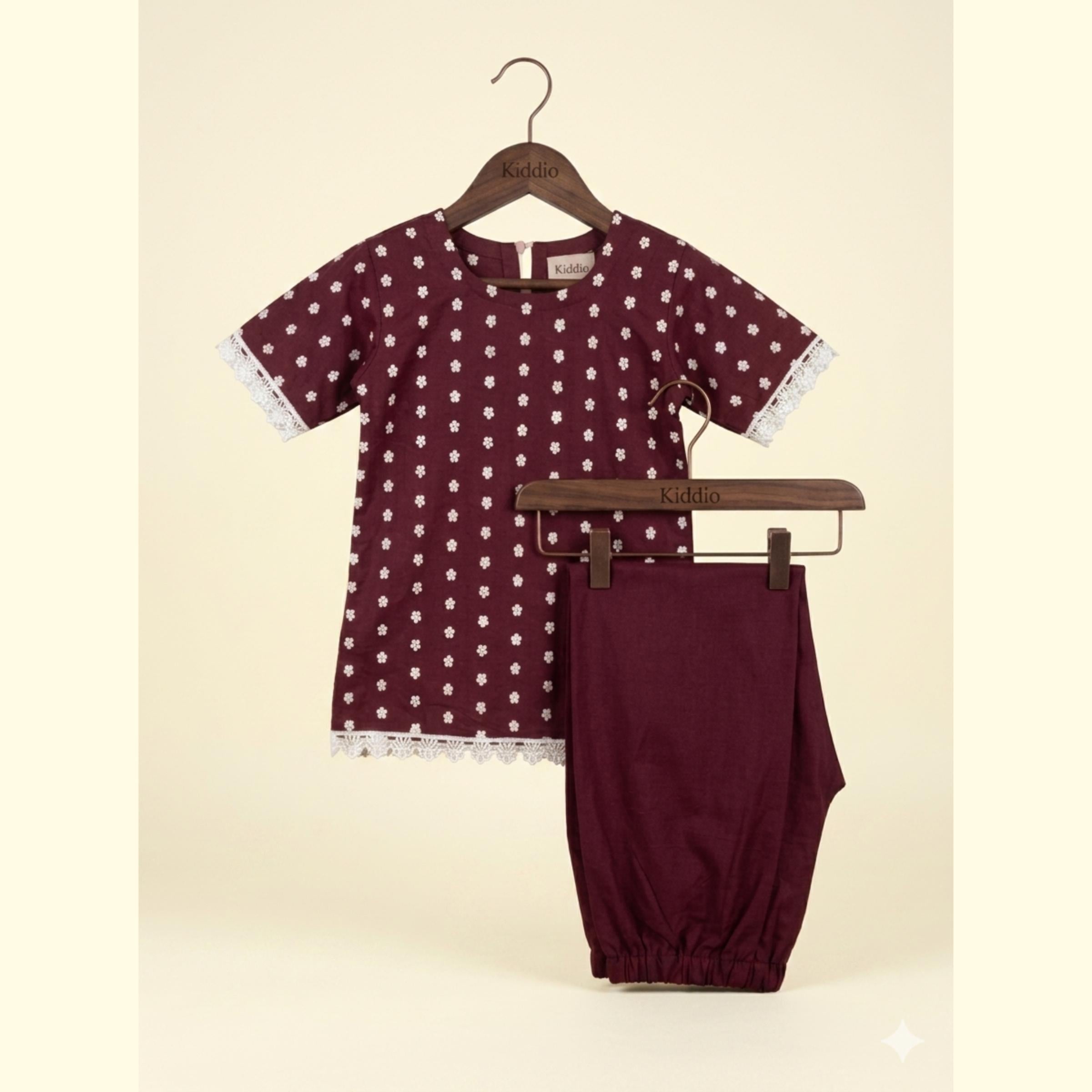 A two-piece baby outfit set consisting of a short-sleeve tunic top and matching pants, displayed on wooden hangers against a light beige background. The set is burgundy with white polka dots and white lace trim.