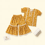 A toddler girl's two-piece clothing set featuring a mustard yellow floral print top and matching wide-leg shorts with white lace trim, accompanied by a small white knit bootie and a KIDDIO brand card on a light cream background.