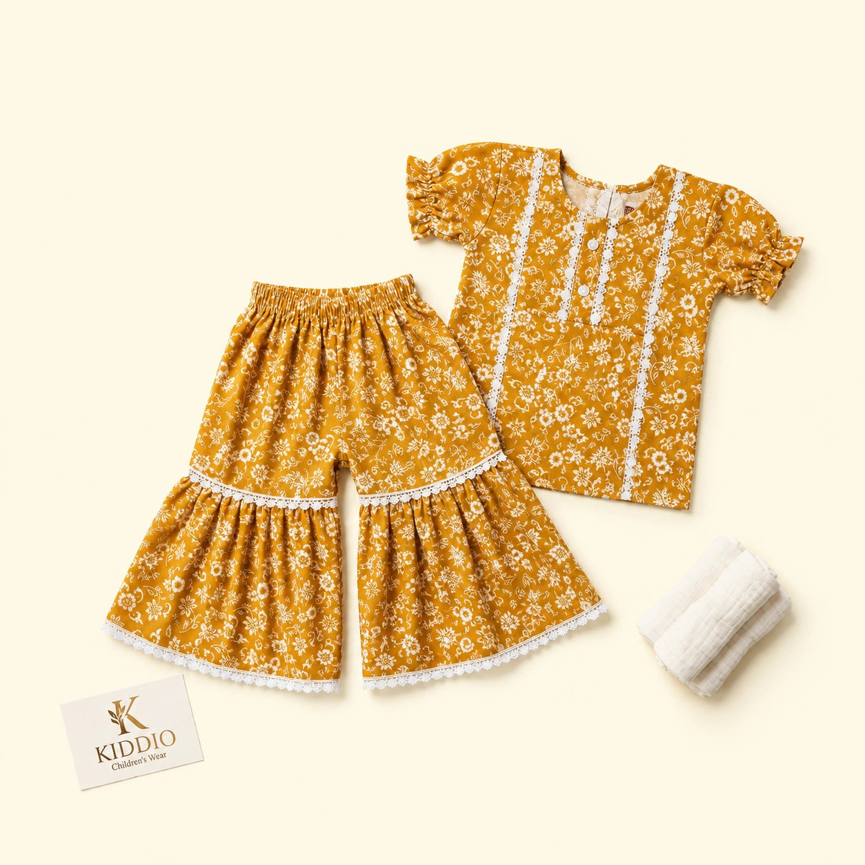 A toddler girl's two-piece clothing set featuring a mustard yellow floral print top and matching wide-leg shorts with white lace trim, accompanied by a small white knit bootie and a KIDDIO brand card on a light cream background.