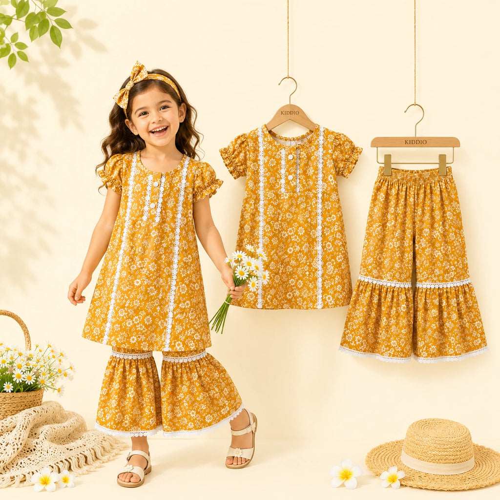 A young girl stands smiling in a matching yellow floral tunic and flared pants set, with the same outfit hanging on hangers beside her. The scene is styled with flowers, a straw hat, and soft cream background, making this a Lifestyle image because a person is present.