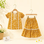 A toddler girl's two-piece outfit set by KIDDIO, featuring a short-sleeve top and matching tiered shorts in a mustard yellow floral print with white lace trim, hanging on wooden hangers against a warm cream background.