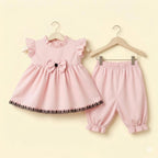 A baby girl's two-piece clothing set in light pink, hanging on wooden hangers against a pale yellow background. The set includes a sleeveless dress with ruffled shoulders, a large bow at the waist, and a decorative black lace hem, paired with matching elastic-waist and ruffled-leg pants.