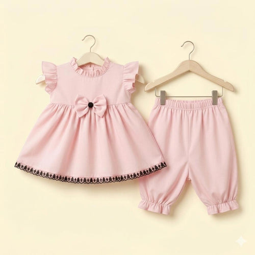 A baby girl's two-piece clothing set in light pink, hanging on wooden hangers against a pale yellow background. The set includes a sleeveless dress with ruffled shoulders, a large bow at the waist, and a decorative black lace hem, paired with matching elastic-waist and ruffled-leg pants.
