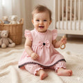 A baby girl sitting on a cream knit blanket in a nursery, wearing a light pink ruffle-sleeve dress with a large bow at the chest, a black embroidered hem, and matching pink ruffled pants. She is holding a wooden teething ring.