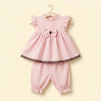 A two-piece baby girl's outfit set in light pink, hanging on a wooden hanger against a pale yellow background. The set includes a ruffled sleeveless top with a bow at the center and a black patterned hem, paired with matching ruffled bloomers.