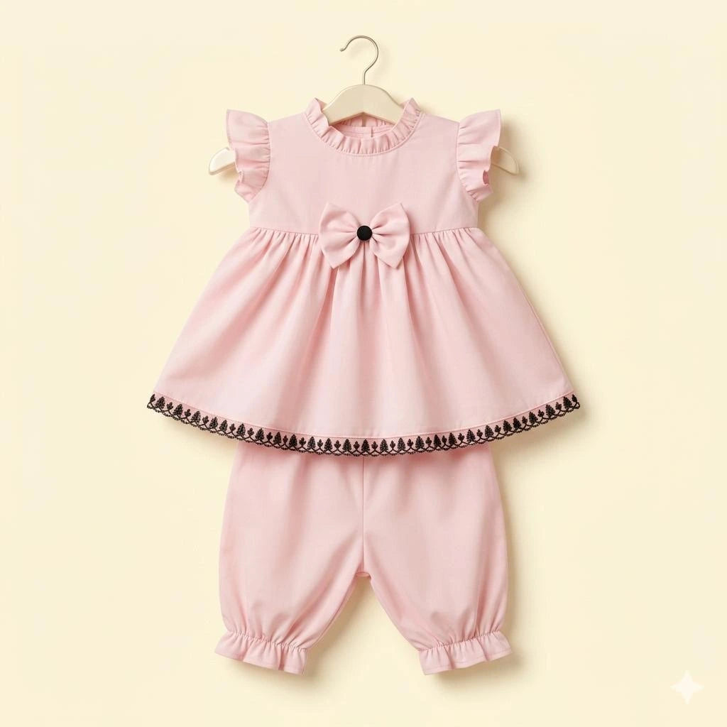 A two-piece baby girl's outfit set in light pink, hanging on a wooden hanger against a pale yellow background. The set includes a ruffled sleeveless top with a bow at the center and a black patterned hem, paired with matching ruffled bloomers.