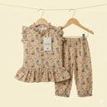 A two-piece children's outfit set on wooden hangers against a solid cream background. The set includes a sleeveless ruffled top and matching pants in a beige base fabric with a playful print of owls, cupcakes, and small animals.